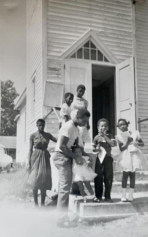 This 1957 photograph was taken in front of the church. The adult male is William H. Daisey Sr. The little girl he is holding is Sarah (Daisey) Minor. The boy next to her is her brother, William H. Daisey Jr. The girl next to him is Fay Collick. The girl at the top of the stairs is Dorothy Riley. The young lady in the dark dress Margie Riley. The remaining girl is unidentified.